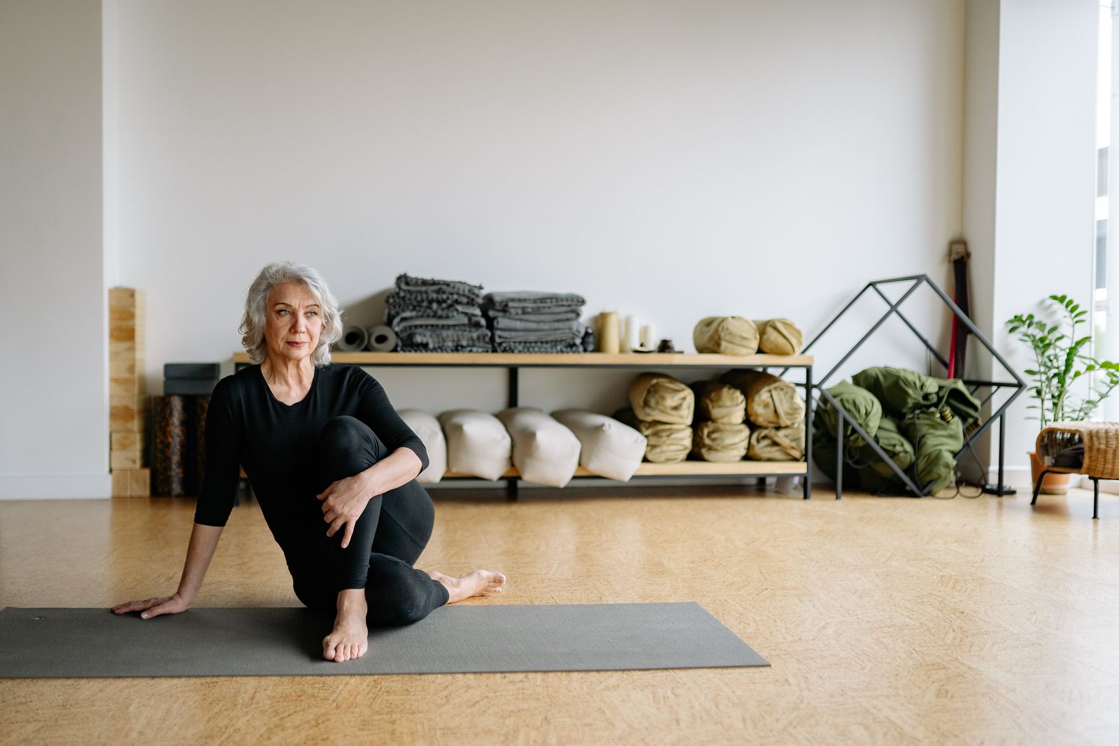 A woman with silver hair seated on a mat by shelves of blankets and bolsters