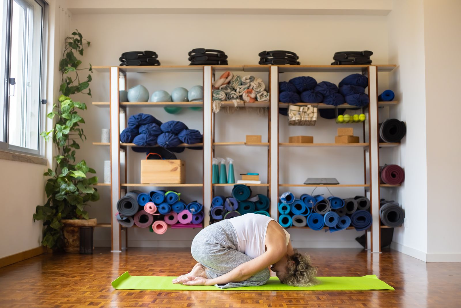 A woman resting in child’s pose on a mat in front of shelves of yoga props