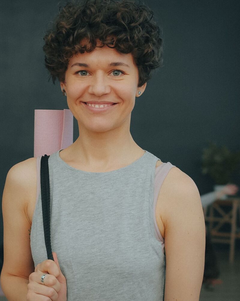 Nora Albrecht, smiling, with a rolled mat over her shoulder in the studio